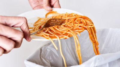 Ett par händer som skrapar ner spaghetti från en tallrik ner i en soptunna.