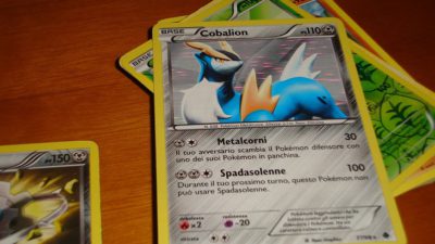 Några pokemonkort ligger i en hög på ett bord. Det översta kortet har gul kant och grå bakgrund. Längst upp är en bild på en blå pokémon med horn. På nedre delen av kortet står text och olika symboler.