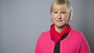 Margot har scarf knuten runt halsen. Hon har runda ringar i öronen och ganska kort hår och lugg. Hon har läppstift och ler lite mot kameran.