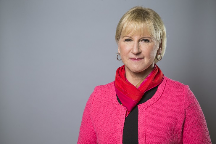 Margot har scarf knuten runt halsen. Hon har runda ringar i öronen och ganska kort hår och lugg. Hon har läppstift och ler lite mot kameran.