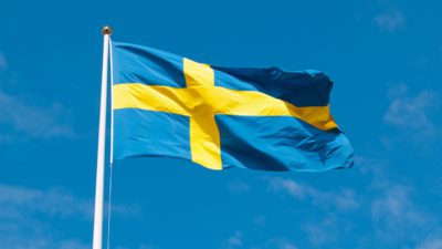 En svensk flagga vajar i vinden på en flaggstång. Solen lyser på flaggan och himlen är blå bakom.