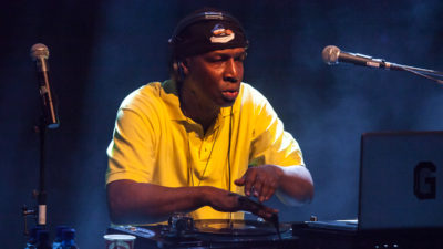 Grandmaster Flash står i ett dj-bås. Han har en backochframvänd keps och stor t-shirt. På bordet framför sig har han dj-utrustning som han spelar med.